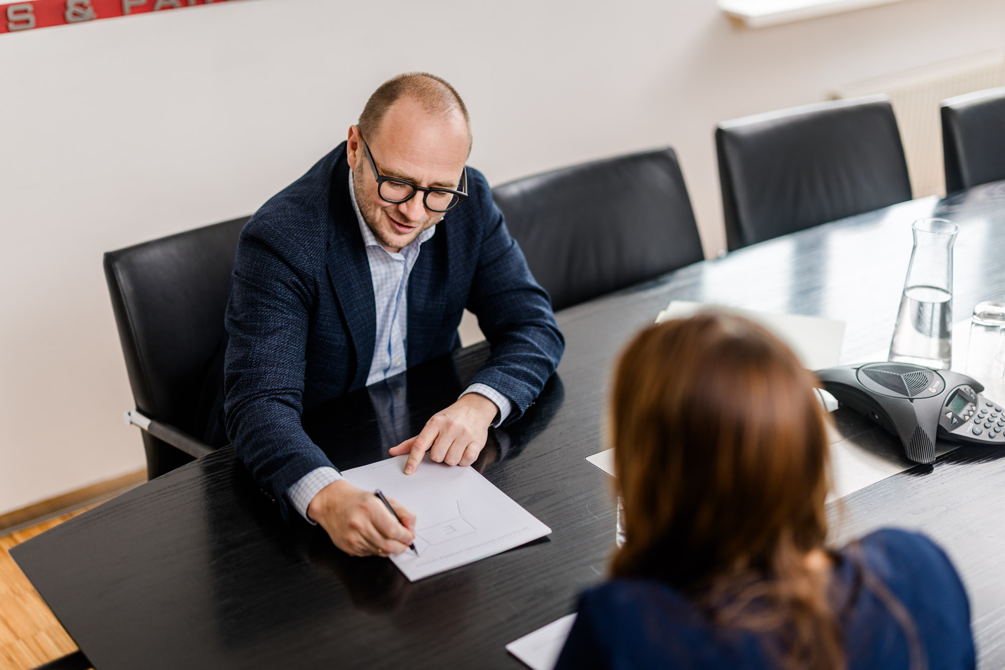 Anwalöt spricht mit einer Dame am Tisch und schreibt etwas auf einen Dokument.