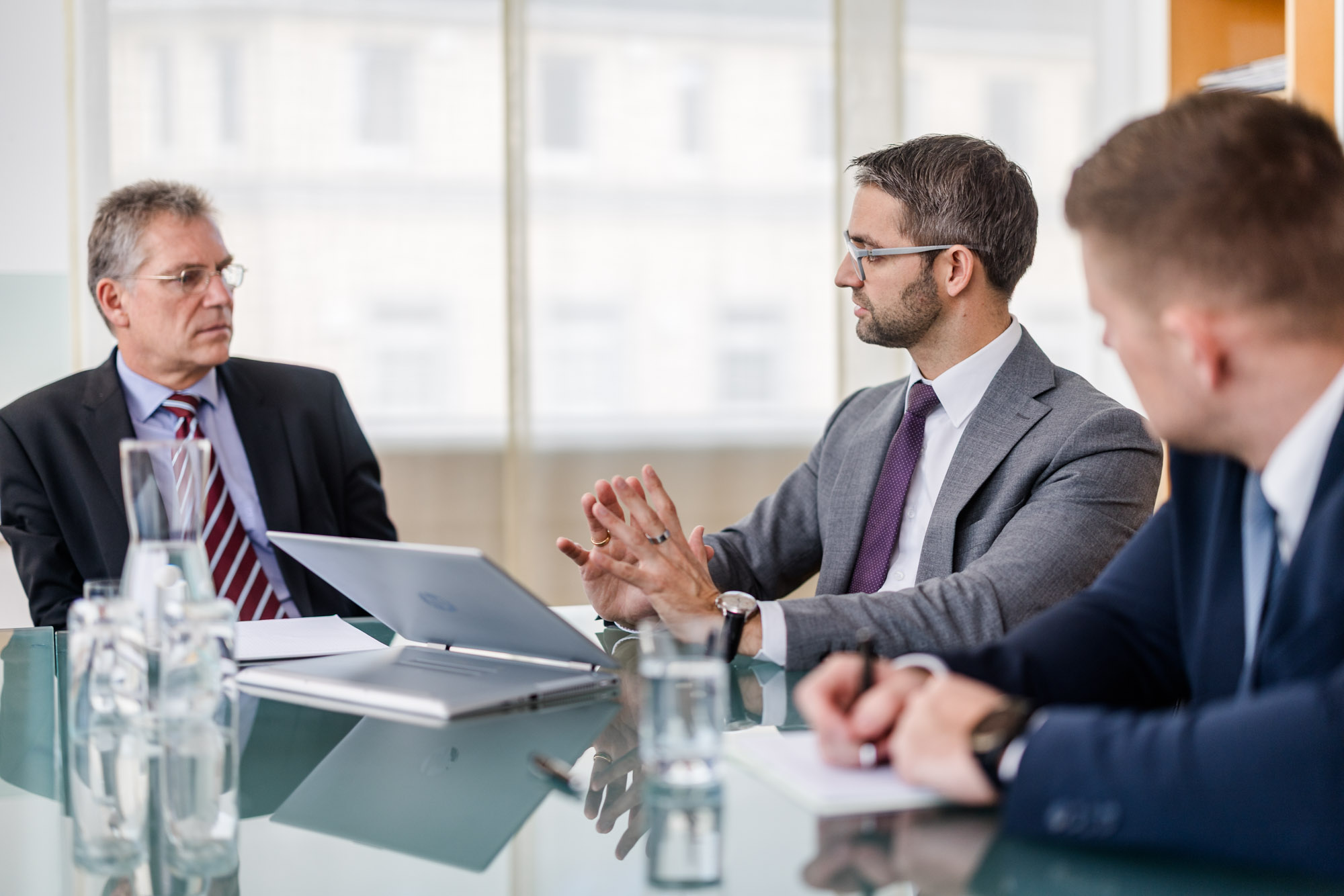 Drei Männer sitzen an einem Tisch mit Laptop und Dokumenten.